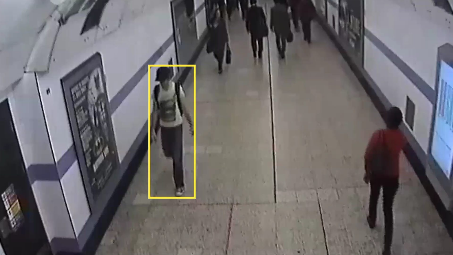 security footage of a person indicated by a yellow tacking rectangle walking through a subway corridor in a baseball hat