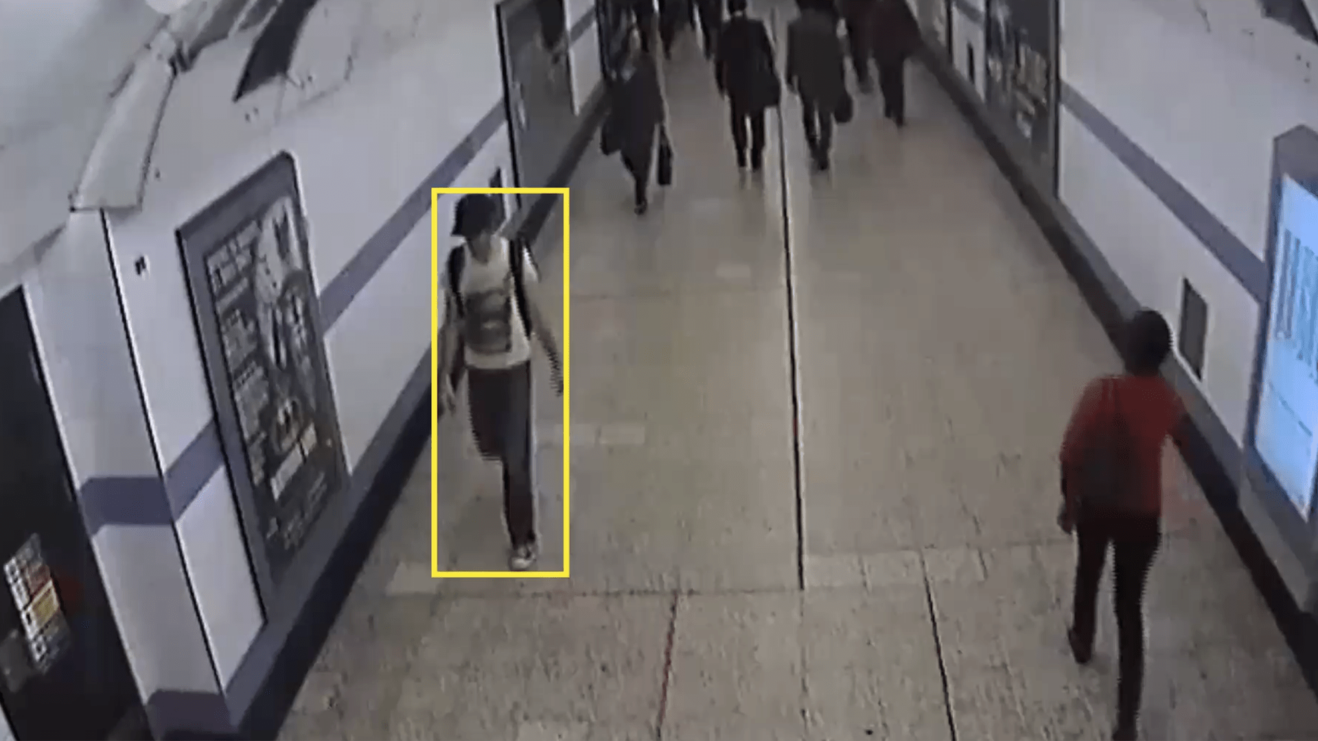 security footage of a person indicated by a yellow tacking rectangle walking through a subway corridor in a baseball hat